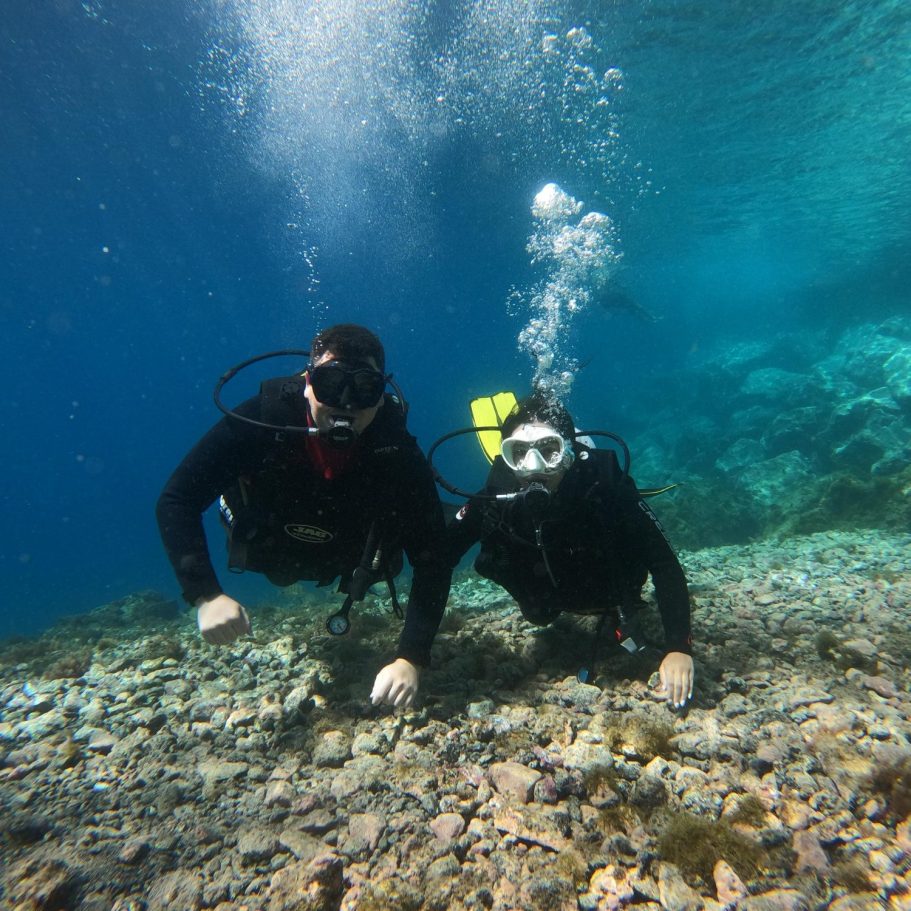 Dos buceadores explorando un fondo marino colorido con corales y burbujas de aire.