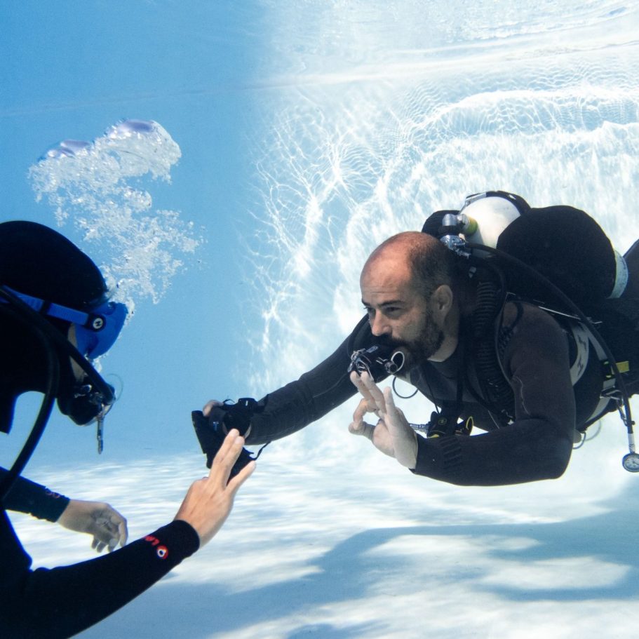 Un buzo interactúa con otro bajo el agua, ambos equipados con equipo de buceo